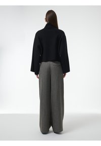 Vrouw met lang haar in een zwarte cropped trui en wijde grijze broek, staand met de rug naar de camera op een effen witte achtergrond.