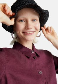 Donna sorridente che indossa grandi orecchini a cerchio e un cappello di paglia scuro, mentre aggiusta il cappello con entrambe le mani, vestita con una camicia bordeaux con bottoni.