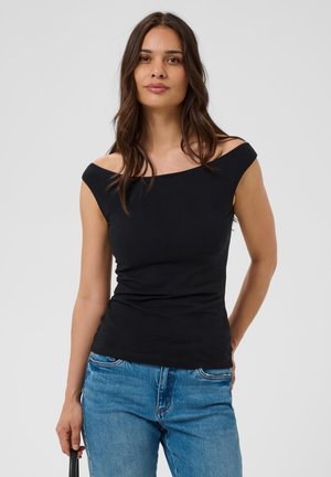 Femme aux longs cheveux foncés portant un haut noir épaules dénudées et un jean bleu, debout devant un fond clair uni.