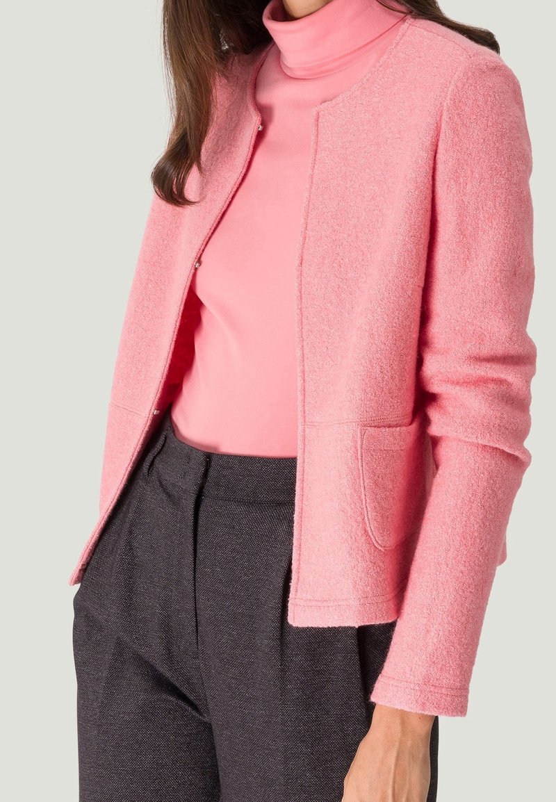 Femme portant une veste texturée rose clair sur un col roulé rose vif, associée à un pantalon taille haute gris foncé.