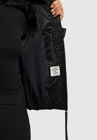 Črna oblazinjena jakna z gladkim, sijočim materialom, z zadrgo na žepu in etiketo znotraj, na kateri piše "Topel objem iz blaga."