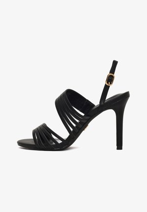 Sandalo nero con tacco alto con diverse sottili fasce sul piede e cinturino alla caviglia con fibbia dorata su sfondo bianco.