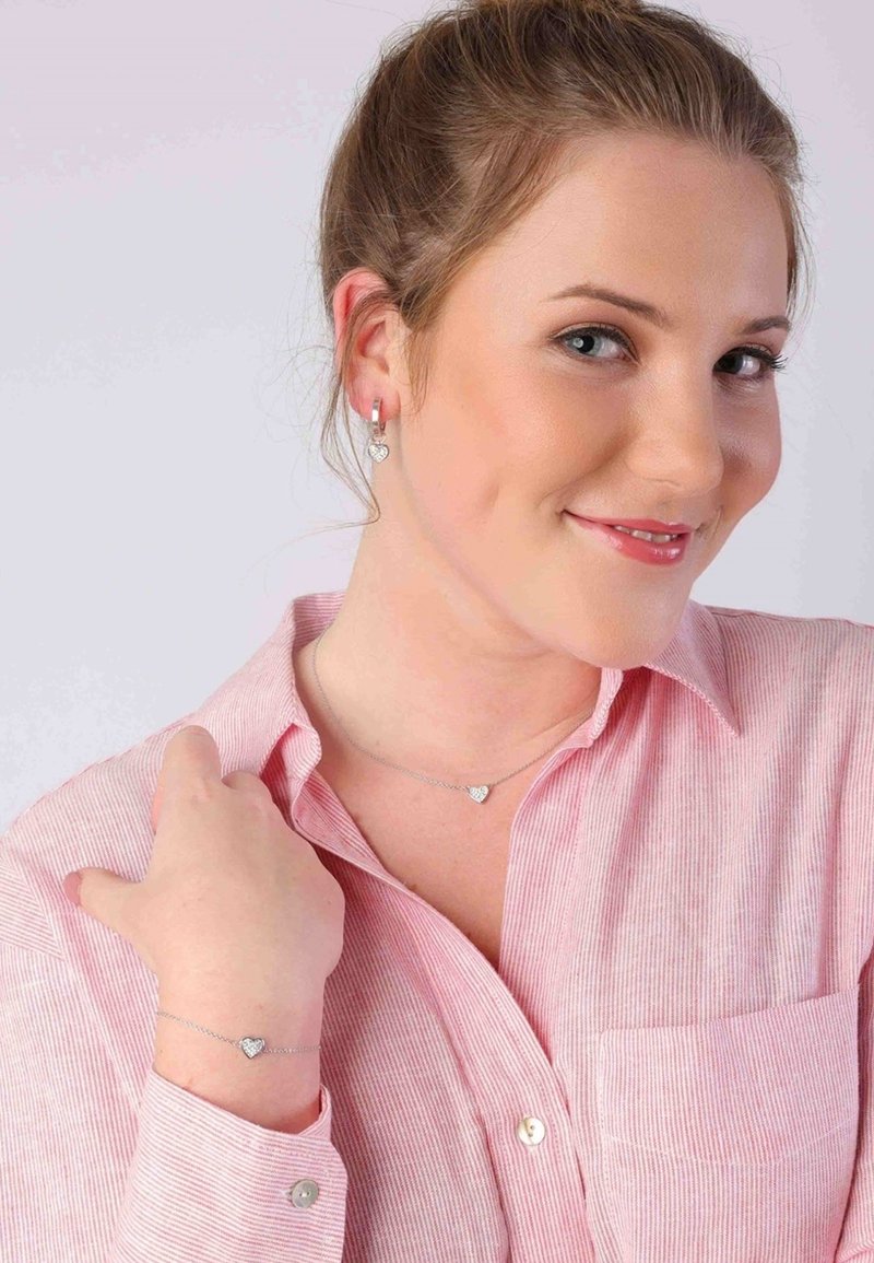 Zilveren hartvormige sieraden omvatten een ketting en een bijpassende armband, gedragen door een model in een roze gestreept shirt.