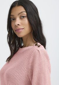 Jeune femme aux cheveux ondulés noirs portant un pull en tricot rose avec deux boutons en laiton sur l'épaule, regardant légèrement sur le côté.