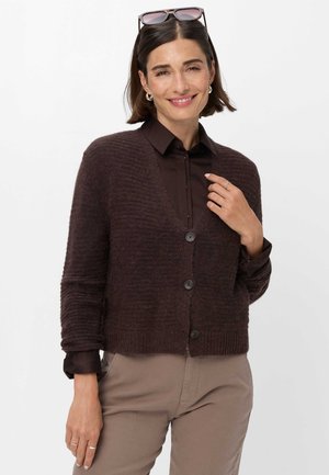 Femme aux cheveux courts et foncés portant des lunettes de soleil sur la tête, un cardigan marron foncé par-dessus une chemise noire, et un pantalon beige, souriant doucement.