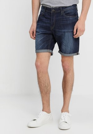 Mann trägt aufgerollte dunkelblaue Denim-Shorts, graues T-Shirt und weiße Turnschuhe, steht vor einem einfachen hellen Hintergrund.