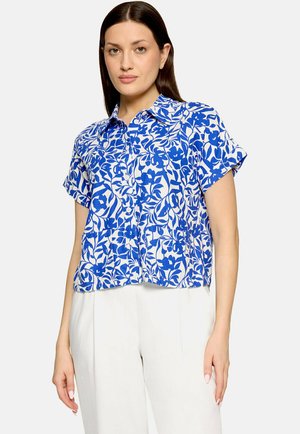 Vrouw draagt een kortärmelig, blauw-wit bloemenpatroon blouse met knopen en witte hooggetailleerde broek op een lichte achtergrond.