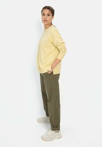 Lichtgele sweatshirt met lange mouwen en een relaxte pasvorm, gecombineerd met olijfgroene broek. Model draagt lichte sneakers met een gestructureerd ontwerp.