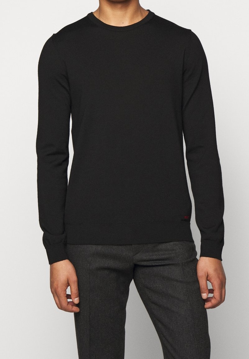 Pull noir à manches longues en tissu côtelé, avec un col rond et un subtil accent rouge à l'ourlet, associé à un pantalon gris foncé.