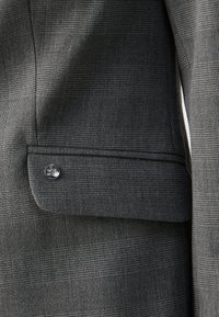 Grijze geruite blazer van textuurstof, met een subtiel ruitpatroon en een zilveren knoop op de voorkant van de zak.