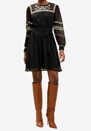 Musta läpikuultava pitkähiha- mekko, jossa valkoiset kirjailut ja vyötäröä korostava leikkaus, yhdistetty polvenkorkuihin ruskeisiin nahkasaappaisiin, joissa on korkeat korot.