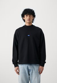 Zwarte sweatshirt met een hoge hals, lange mouwen en een blauwe rechthoekige logo op de borst. Draag het met lichtblauwe jeans.