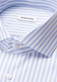 Gros plan sur une chemise blanche pliée avec des rayures en chevrons bleues, poignet boutonné et étiquette Seidensticker cousue à l'intérieur du col.