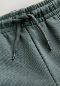 Green textured fabric of sweatpants featuring an elastic waistband and a drawstring with black tips, showcasing a close-up view of the design.