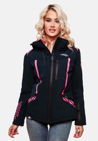 Blonde woman wearing a black and pink Mariko Mountain zip-up jacket with hood and blue jeans, standing against a white background.