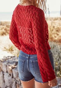 Pull en maille rouge avec un motif en zigzag, associé à un short en denim bleu. Le pull comporte des manches longues et un design texturé à tissage ouvert.