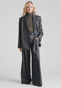 Szary, dopasowany blezer z podwójnym rzędem guzików, zestawiony z szerokimi, szarymi spodniami. Pod spodem oliwkowy golf. Gładka, strukturalna tkanina.