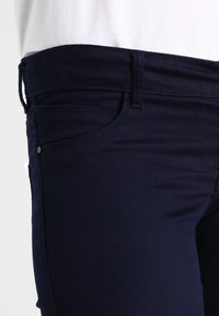 Námořnické modré skinny džíny vyrobené z elastického materiálu s hladkým povrchem, s klasickým pětizakládovým designem a minimálními detaily šití.