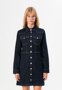 Kvinne iført en mørk jeansskjorte med lange ermer, to brystlommer og kontrasterende beige sømmer mot en helt hvit bakgrunn.