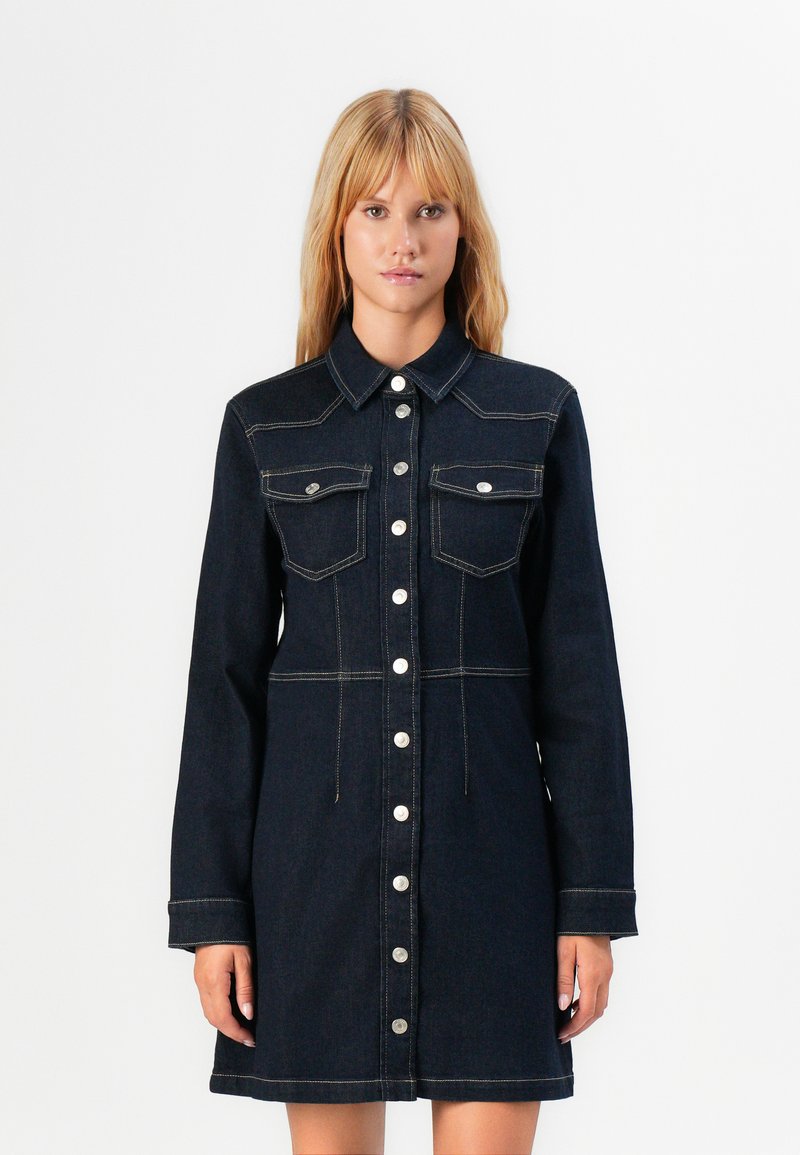 Kvinne iført en mørk jeansskjorte med lange ermer, to brystlommer og kontrasterende beige sømmer mot en helt hvit bakgrunn.