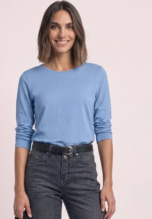Smilende kvinde med skulderlangt brunt hår iført en lyseblå langærmet skjorte og mørkegrå jeans med sort bælte.