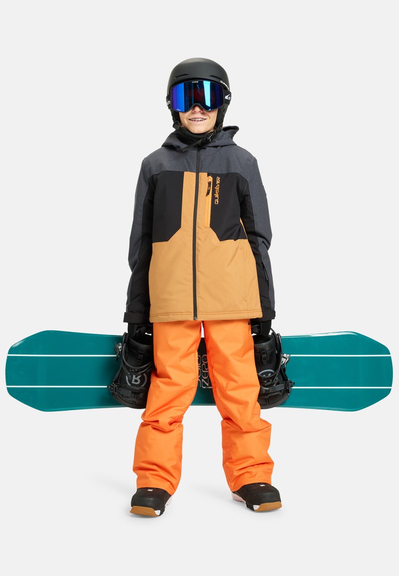 Snowboarder gekleed in een zwart en oranje waterdichte jas, heldere oranje broek, en met een zwarte helm en goggles, die een turquoise snowboard vasthoudt.