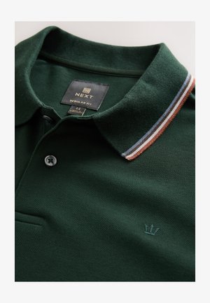Polo en coton vert foncé avec une finition texturée, doté d'un col à rayures marron, bleu clair et blanc, ainsi que d'un petit logo couronne.