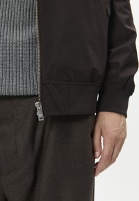 Must bomberjakk ribadeeritud halli kampsuni all. Silmad hõbedast tõmblukku, elastsed mansetid ja tekstuuriga disain. Pruunid püksid.