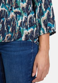 Blouse à motifs dans des teintes de bleu, vert et beige, dotée d'une texture douce et de manches trois-quarts. Détail de logo notable à l'ourlet.