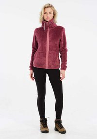 Giacca in pile bordeaux con collo alto e zip frontale, abbinata a leggins neri e scarponi da trekking marroni, su uno sfondo neutro.
