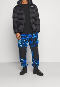 Chaqueta de plumas negra con capucha, diseño grueso; combinada con pantalones de tela tipo tie-dye en azul y negro, con parches de tela negra, y zapatos negros.