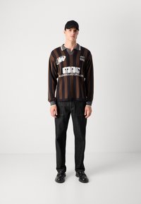 Uomo che indossa un berretto nero, una camicia a maniche lunghe a strisce marroni e nere con testo bianco, pantaloni neri e scarpe nere, in piedi di fronte a un muro semplice.