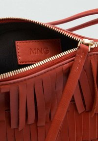 Sac en cuir rouge avec des franges, fermeture éclair dorée et intérieur noir. Logo MNG visible sur une petite étiquette en cuir à l'intérieur.