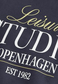 Sweatshirt navy avec texte blanc et doré : "Leisure STUDIOS Copenhague EST 1982." Texture lisse, design décontracté et branding bien visible.