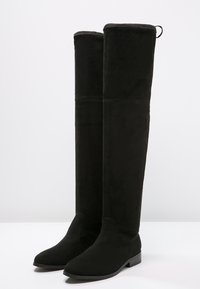 Botas de ante negras hasta la rodilla con una textura suave, puntera afilada y tacón plano. Presentan una costura vertical y una lengüeta en la parte superior.