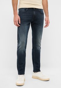 Jeans de mezclilla azul oscuro con un ajuste slim, un ligero desteñido y un diseño de cinco bolsillos. Combinados con zapatillas blancas, mostrados sobre un fondo neutro.