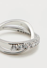 Ασημένιο δαχτυλίδι crossover με δύο μπάντες, μία από τις οποίες είναι διακοσμημένη με μικρές στρογγυλές διαφανείς πέτρες κατά μήκος της καμπύλης.