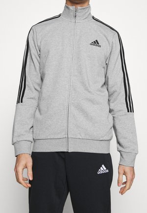 Mand iført en grå Adidas lynlåstrøje med sorte striber på ærmerne og sorte Adidas bukser med hvidt logo.