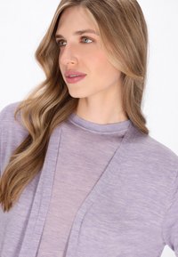 Paarse lichtgewicht cardigan met een gestructureerde afwerking, open voorkant en geribbelde halslijn, gelaagd over een zachte paarse top.