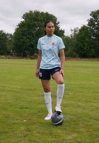 Kvinnefotballspiller står på gresset, iført en lys blå geometrisk mønstret trøye med et merke og mørke shorts, hvite strømper og fotballsko.