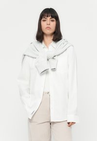 Donna con i capelli scuri di lunghezza media che indossa una camicia bianca con bottoni, pantaloni beige chiaro e un maglione grigio chiaro appoggiato sulle spalle.