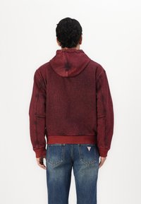 Rød sweatshirt med hætte, der viser en falmet tekstur og synlige syninger. Båret med blå jeans med et karakteristisk lomme-design.