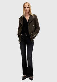 Giacca da biker in suede verde oliva con zip, top nero, jeans neri svasati e scarpe nere con dettagli dorati. Texture liscia e vestibilità sartoriale.