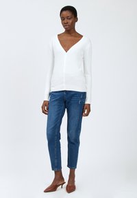 Weißer, langärmeliger Cardigan mit tiefem V-Ausschnitt und Knopfverschluss, kombiniert mit blauen Jeans und braunen, hohen Mules. Einheitlicher Hintergrund.