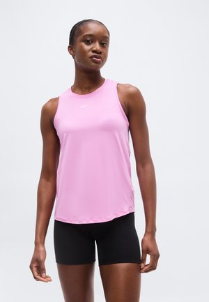 Naine, kes kannab heleroosat varrukateta sportlikku topi ja musti lühikesi pükse, seisab heleda halli tausta ees.