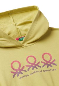 Sudadera amarilla con un logo y texto en rosa de United Colors of Benetton en el pecho, mostrando la costura interior de la capucha.