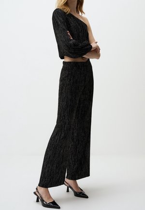 Vrouw draagt zwarte, gestructureerde wijde broekspijpen en een zwarte top met lange mouwen en één blote schouder, met zwarte puntige hakken.