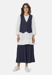 Marineblaues Denim-Weste mit einem eingekerbten Kragen, drei Knöpfen, über einer weißen Tunika und einem marineblauen Rock getragen, vervollständigt mit weißen Sneakers.