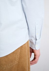 Light blue collared shirt with long sleeves, featuring buttoned cuffs. The shirt is tucked into brown corduroy pants with a textured finish.