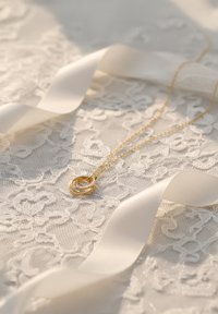 Guldhalskæde med vedhæng af sammenflettede ringe sat på hvidt blondebaggrund, ledsaget af blød hvid satinbånd. Delikat kædedesign.